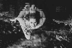 Žena ve vodě – černobílá fotografie, žena v letních šatech leží na zádech ve vodě, její obraz se zrcadlí na vodní hladině; fotografie je obrácená (takže hladina je nahoře)
