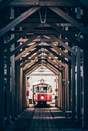 Tramvaj – barevná fotografie, historická tramvaj v dálce v depu, pohled je rámován dřevěným trámovím vztyčeným nad kolejemi