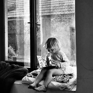 Chvilka s knihou na okně – černobílá fotografie, malý chlapec sedí na dece na okně starého domu a čte si v knížce